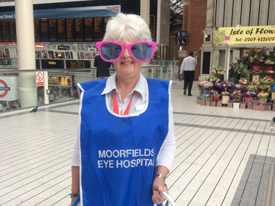 Picture of a collector in a blue tabard wearing large pink sunglasses at Liverpool Street Station.