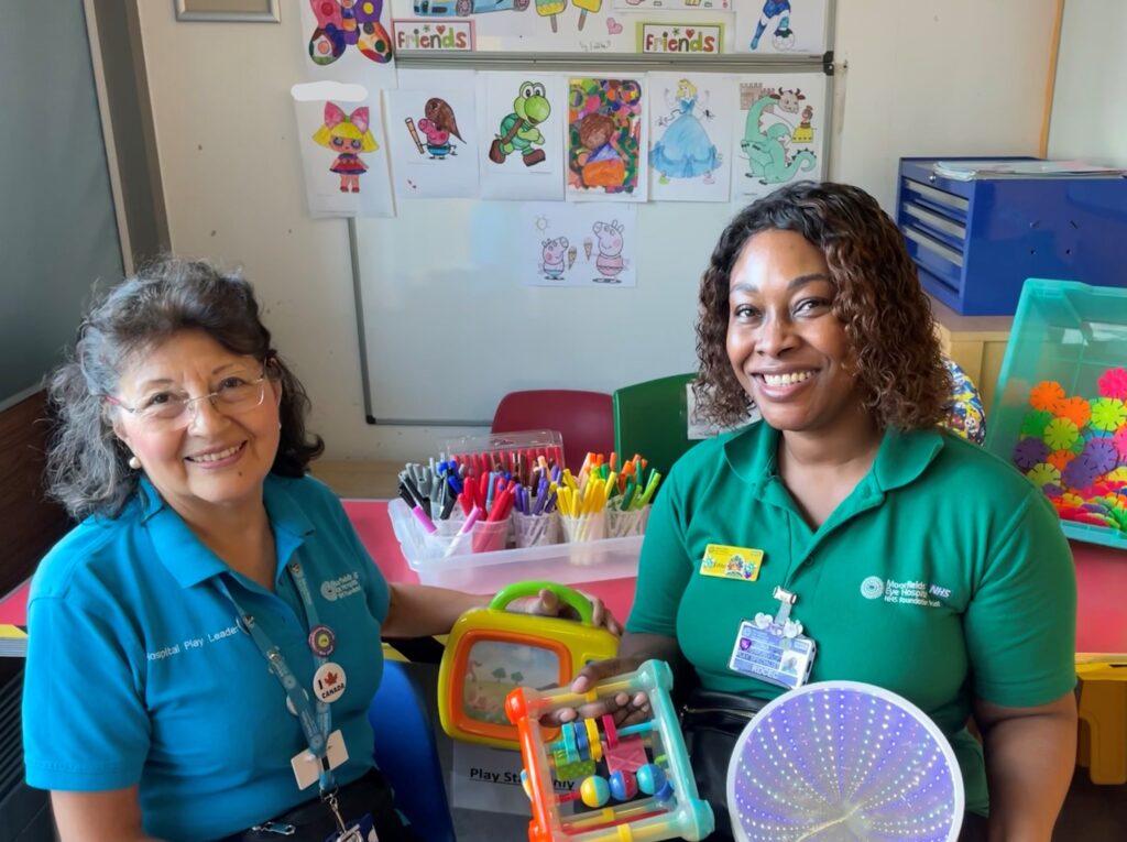 A photograph of Estela, a play leader, and Edna, a play specialist. They are sitting and smiling, surrounded by toys and colouring pages.