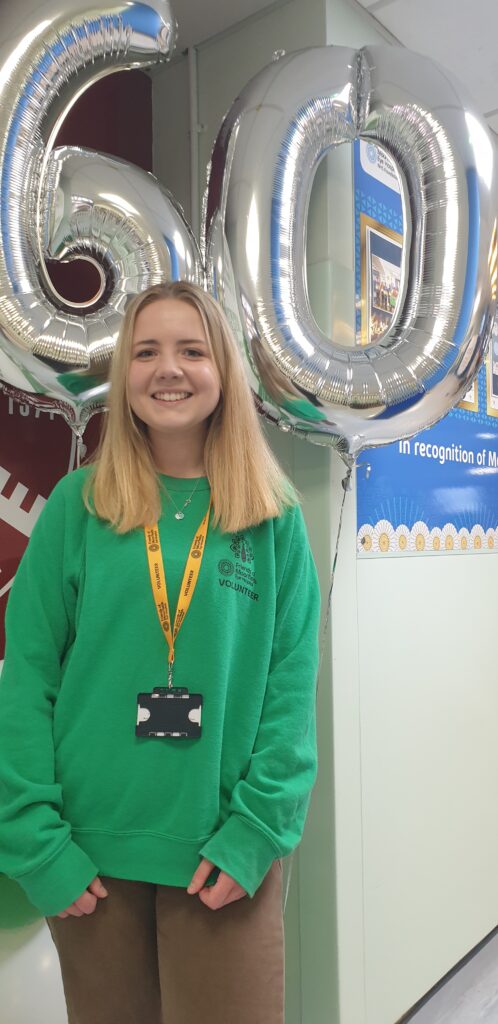 Sophie, a volunteer wearing a green jumper, stands smiling in front of silver balloons in the shape of the number 60.