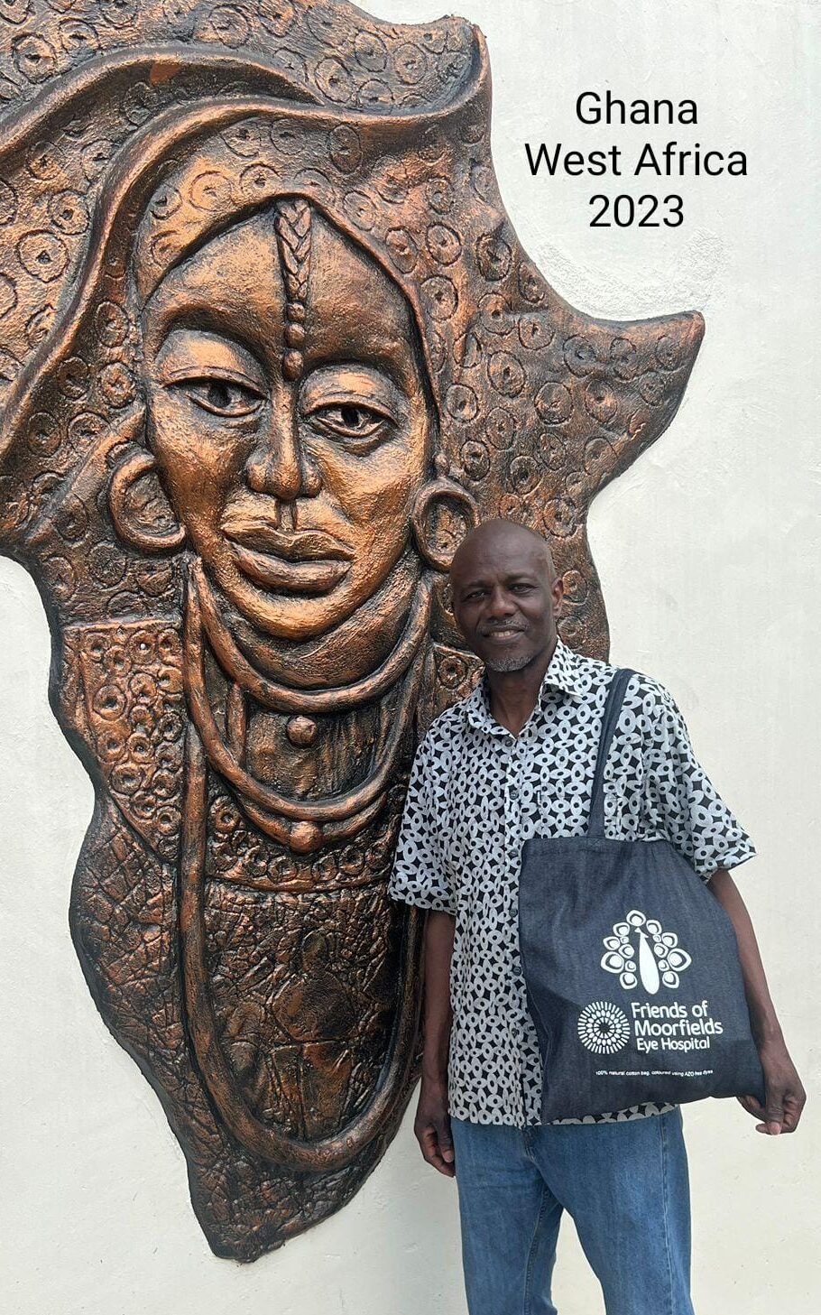 A photo of Robert posing with a Friends of Moorfields tote bag, in front of an artwork in Ghana.