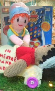 Two knitted dolls. One is lying on a bed. The other is dressed in scrubs and holding the first doll's hand.