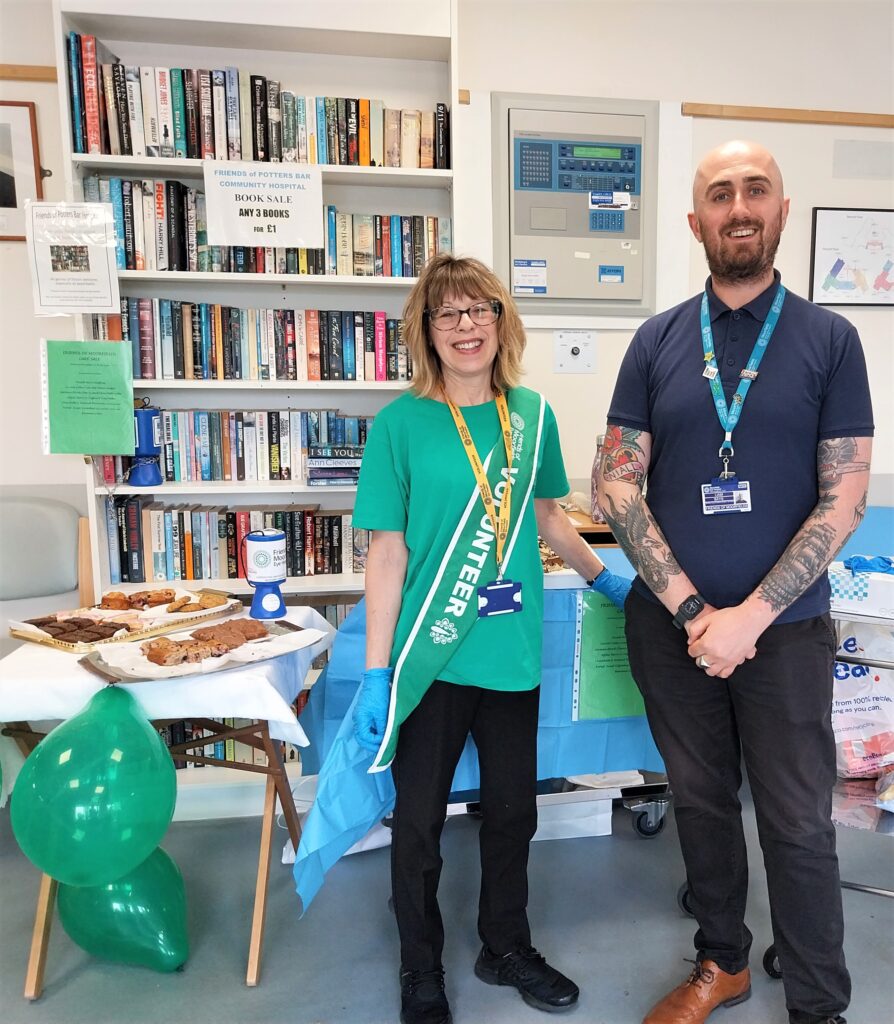 A photo of Jackie, in a green volunteer uniform, and Liam, standing beside a table of cakes and a collecting tin.
