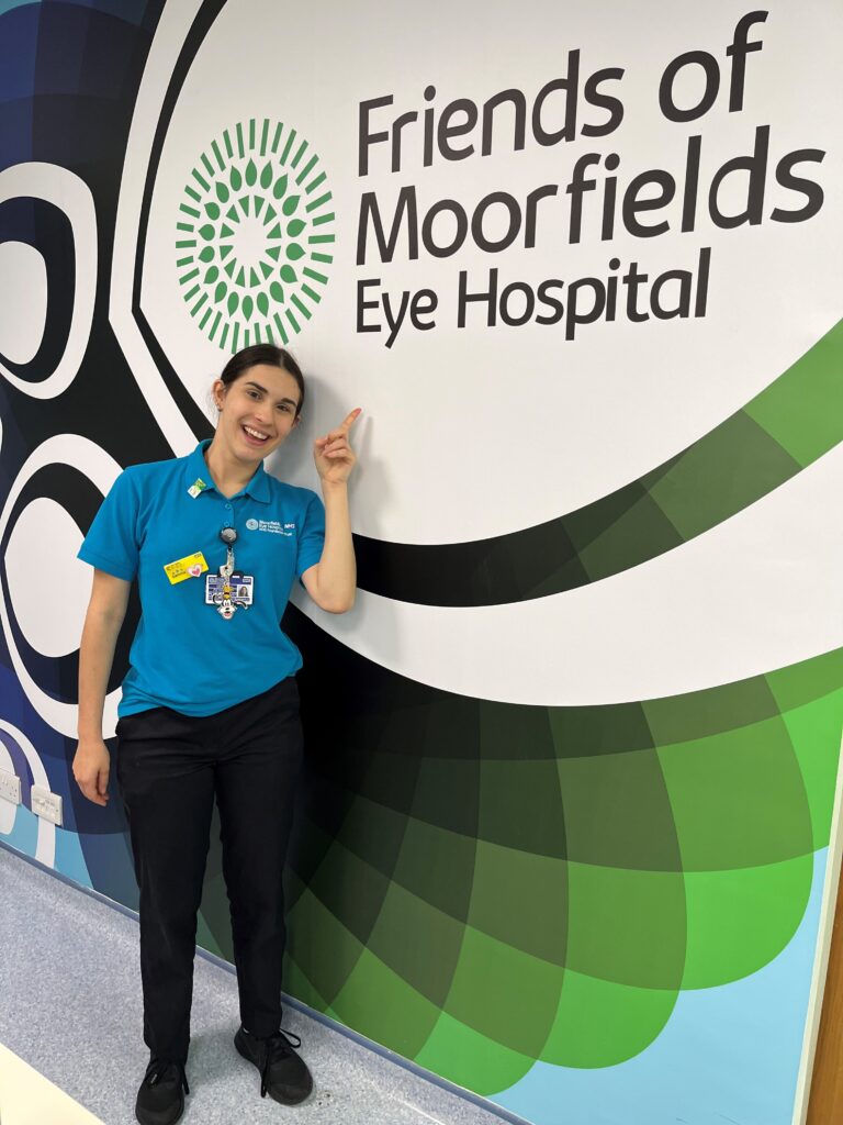 Gemma, dressed in her Health Play Specialist uniform t-shirt, smiles as she points to a large Friends of Moorfields logo on the wall.