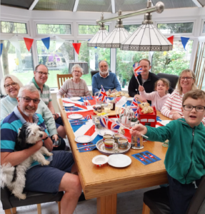 A photo of a large family group of multiple generations enjoying afternoon tea in a conservatory decorated with Union Jacks and red, white and blue bunting.