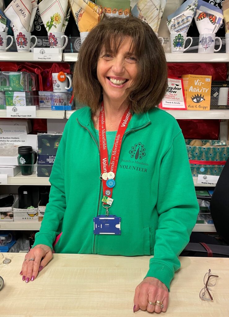 Eve behind the counter of the Friends shop. She is smiling and wearing a green Friends of Moorfields volunteer jacket.