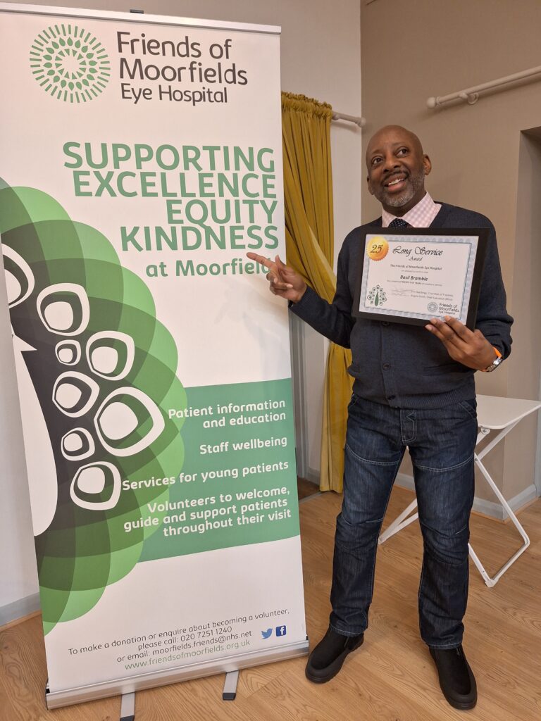 Basil stands with a certificate marking 25 years of service, and points to a Friends of Moorfields banner that says "Supporting excellence, equity, kindness at Moorfields".