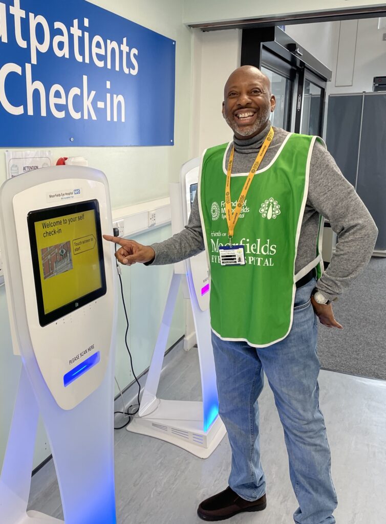 Basil, wearing a green volunteer tabard, stands smiling beside a check-in machine at the entrance to Moorfields City Road.