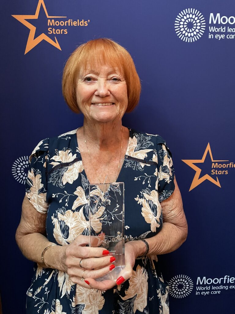 Brenda smiles, holding her award, in front of a Moorfields Stars backdrop