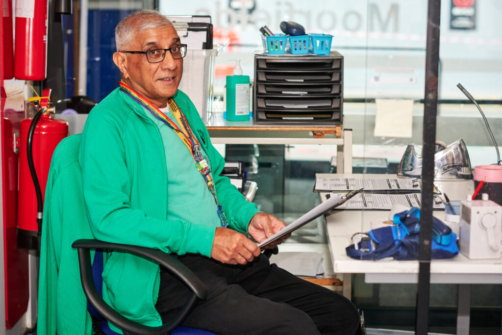 Kiran, a volunteer, sits smiling with a clipboard at a desk near the entrance of A&E.