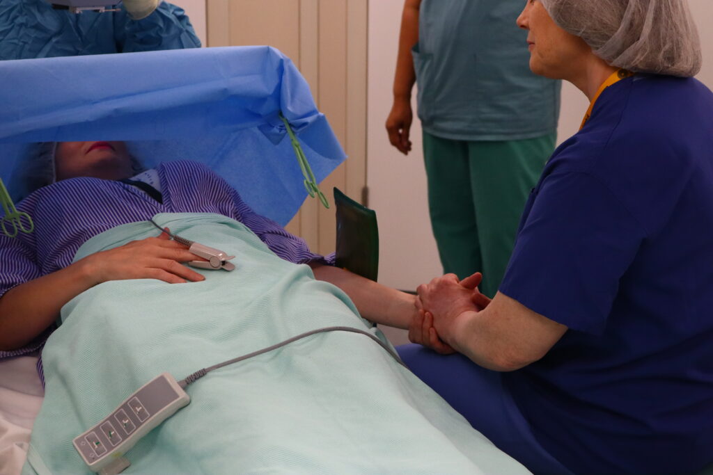 A volunteer in scrubs holds the hand of a patient lying on a bed, as in the background staff prepare for a procedure.