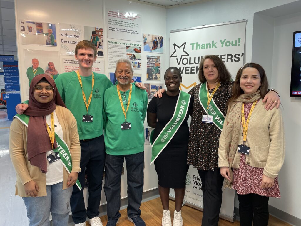 A group of volunteers in green uniform pose smiling together in front of a Volunteers' Week display.