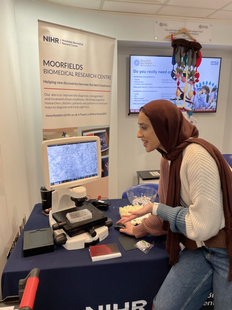 A researcher talks, gesturing towards a screen showing a close-up view of a microscope slide.