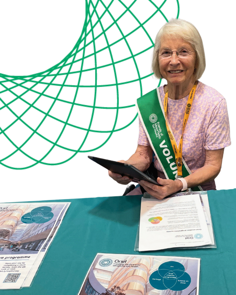 A smiling volunteer sits at a table holding a clipboard. On the table are leaflets with pictures of the new centre.